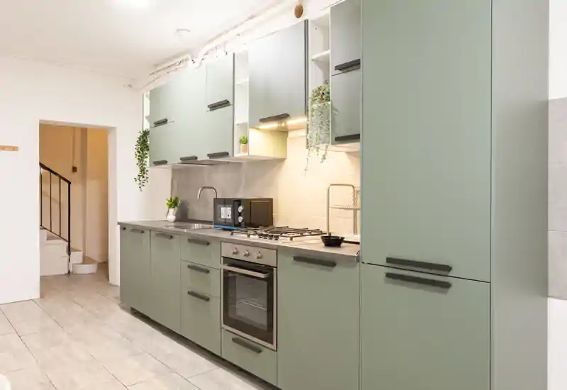 Cucina moderna con mobili verde salvia, forno integrato e piano cottura a gas, microonde sul piano e piccole piante decorative. Spazio luminoso e ordinato ideale per annunci di appartamenti.