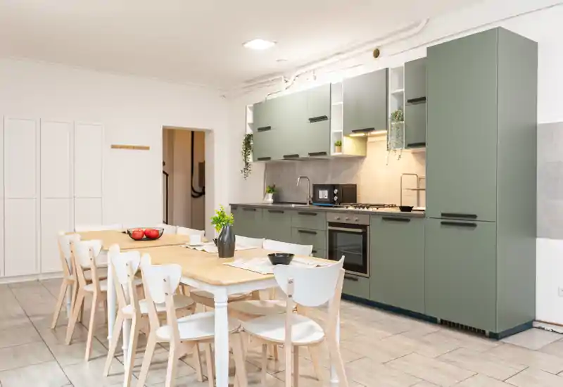 Cucina moderna e luminosa con mobili verdi e un grande tavolo da pranzo in legno per otto persone, arredata con piante e stoviglie — spazio cucina-sala da pranzo aperto e funzionale.