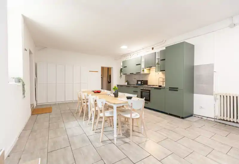 Ampia zona pranzo open space con un lungo tavolo in legno e sedute per più ospiti, adiacente a una cucina moderna verde — luminosa, essenziale e perfetta per pasti in compagnia.