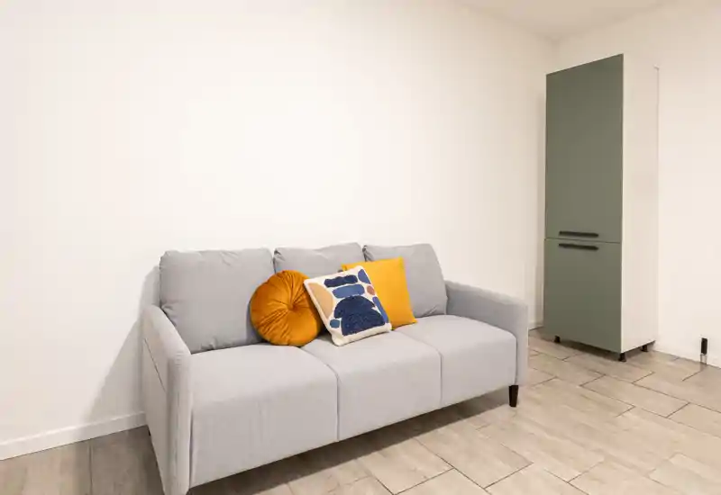 Zona living contemporanea con divano grigio chiaro a tre posti e cuscini colorati, parete bianca pulita e pavimento piastrellato — luminosa, minimale e ben curata per un annuncio.