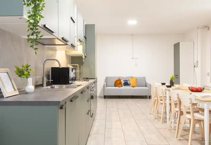 Cucina open-space moderna con mobili verde salvia, lavello integrato e tavolo da pranzo; un piccolo divano sullo sfondo e piante decorative rendono l'ambiente accogliente e funzionale.