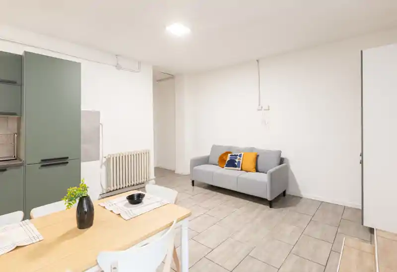 Zona giorno luminosa e minimal con un piccolo divano grigio con cuscini colorati, un tavolo in legno in primo piano e parte di una kitchenette visibile a sinistra.
