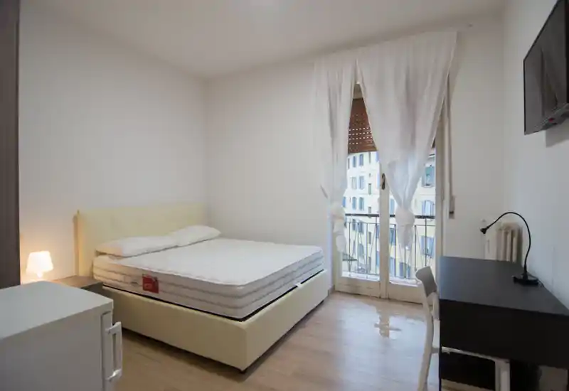 Dormitorio luminoso y minimalista con cama doble, lámpara de noche y escritorio junto a la puerta del balcón que aporta luz natural. Líneas limpias, tonos neutros y ropa de cama sencilla crean un espacio ordenado y acogedor, ideal para alquileres temporales.