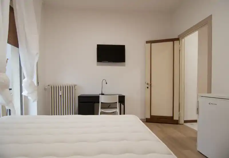 Dormitorio luminoso y minimalista con el colchón en primer plano, un pequeño escritorio con televisor en la pared y una ventana-balcon que aporta luz natural.