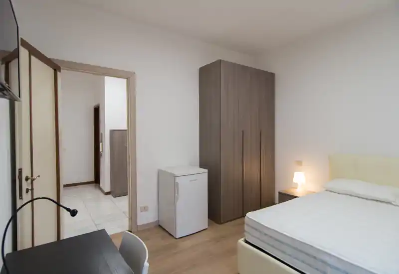 Dormitorio luminoso y sencillo con cama doble, mesilla con lámpara, armario y pequeño frigorífico. Tonos neutros y líneas limpias que crean un ambiente tranquilo, ideal para estancias cortas.