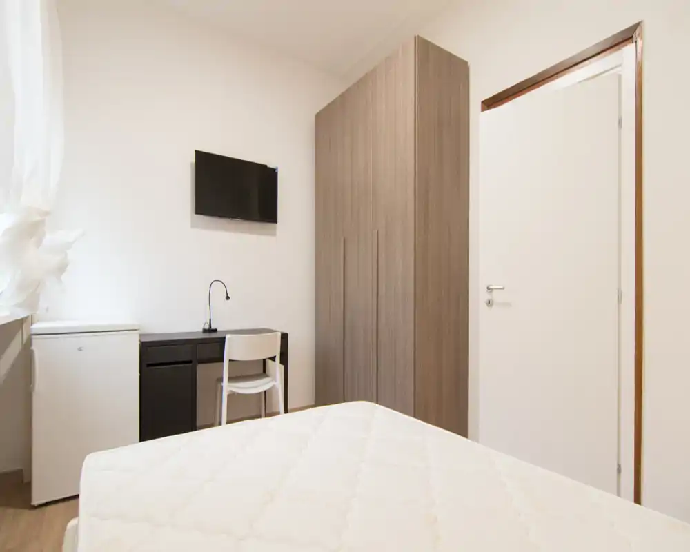 Dormitorio luminoso y minimalista con colchón en primer plano, armario, pequeño escritorio y TV en la pared: distribución sencilla y funcional, ideal para estancias cortas.