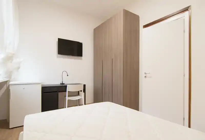 Dormitorio luminoso y minimalista con colchón en primer plano, armario, pequeño escritorio y TV en la pared: distribución sencilla y funcional, ideal para estancias cortas.