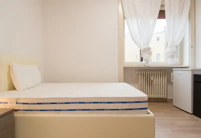 Dormitorio sencillo y ordenado con colchón y almohada visibles, luz natural suave desde la ventana y mobiliario mínimo para un aspecto limpio y moderno.