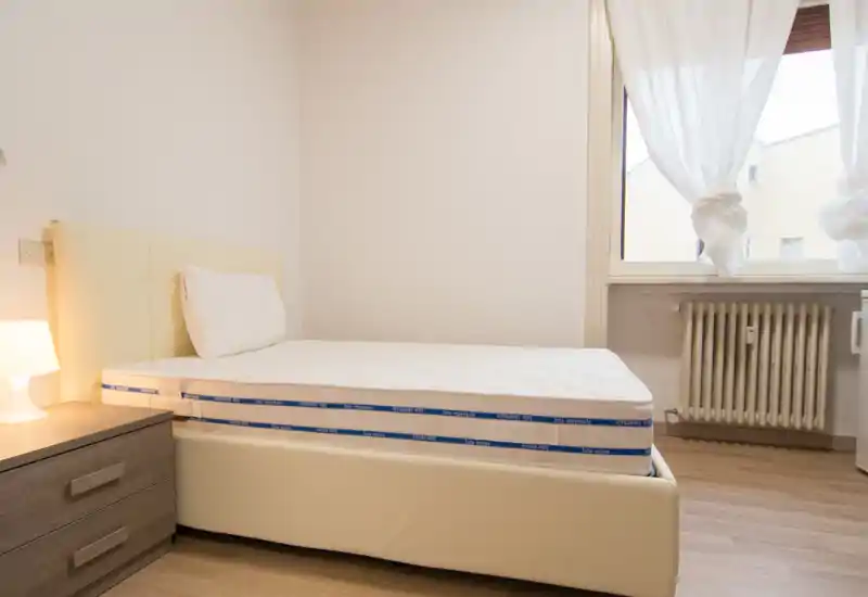 Dormitorio luminoso y sencillo con colchón limpio sobre una cama baja tapizada, mesita con lámpara y ventana con cortinas translúcidas que aporta luz natural.