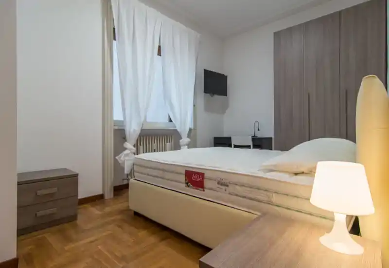 Camera da letto moderna e luminosa con letto matrimoniale, lampada da comodino e armadio; finestra con tende bianche e piccolo scrittoio con TV a parete.