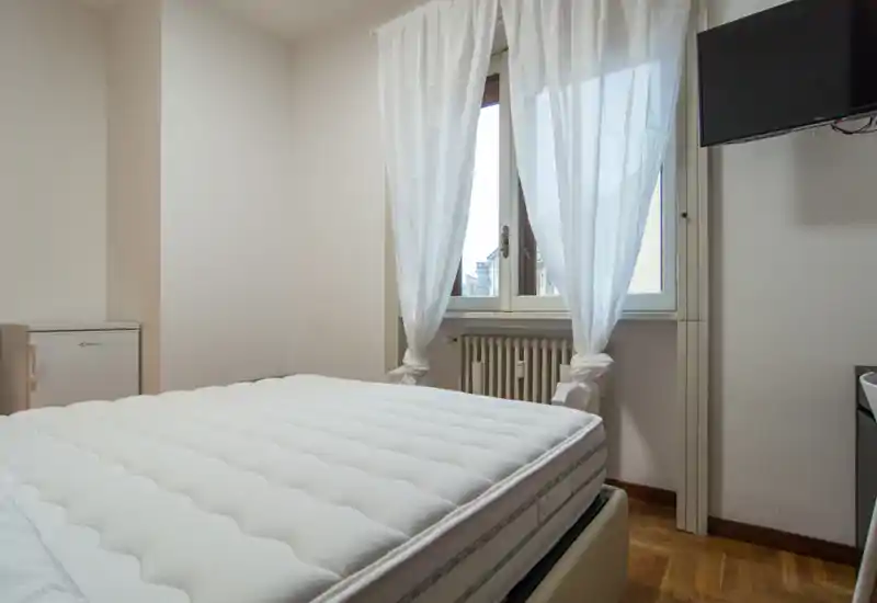 Dormitorio luminoso y de estilo sencillo con colchón amplio, cortinas blancas y TV en la pared; la luz natural crea un ambiente limpio y aireado.