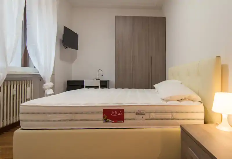 Dormitorio luminoso de estilo sencillo con colchón grande visible, cabecero acolchado, mesita con lámpara y armario; la luz natural crea un ambiente acogedor.