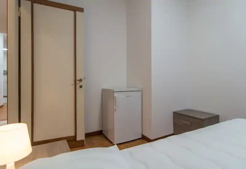 Angolo camera pulito con materasso visibile, comodino, lampada e un piccolo frigorifero. Arredamento neutro e pavimento in legno creano un'area notte semplice e funzionale.