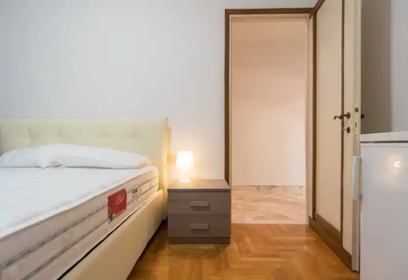 Dormitorio luminoso y minimalista con una gran cama tapizada, mesita de noche y lámpara cálida; suelo de parquet y paredes neutras que transmiten calma y modernidad.