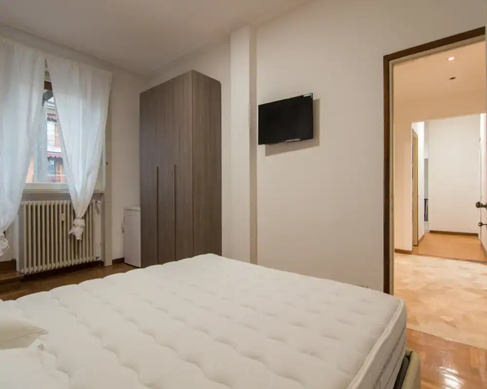 Bright, simply furnished bedroom with a large visible mattress, wardrobe and wall-mounted TV. Soft natural light from the window and neutral decor create a calm atmosphere.