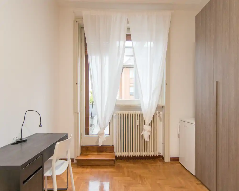 Bright, simply furnished bedroom-sized space with large window/door, white curtains, a desk and wardrobe — ideal for a single occupant focusing on natural light and clean lines.