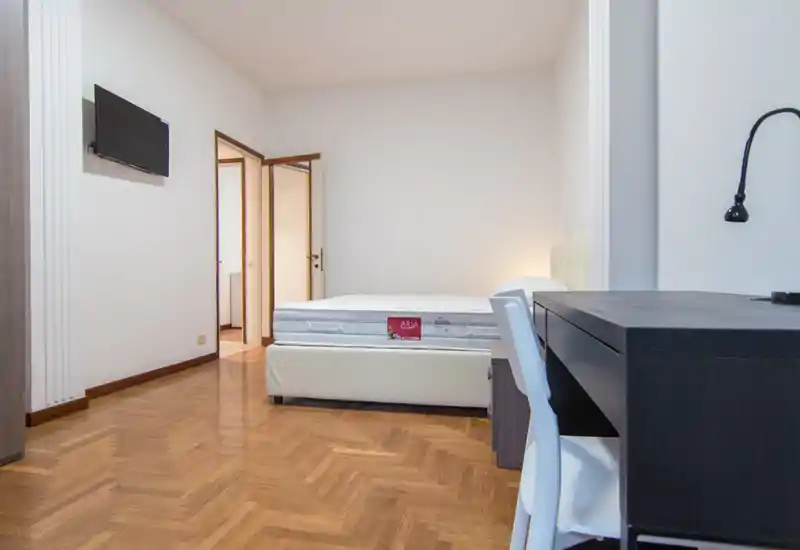 Bright, minimal bedroom with a visible double bed, parquet flooring and a work desk in the foreground. Clean, modern lines and soft ambient lighting create a calm atmosphere ideal for rental listings.