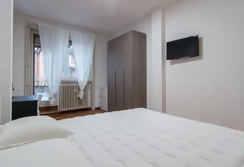 Bright and neat bedroom with a large bed in the foreground, a wardrobe, window with sheer curtains and a wall-mounted TV — comfortable and simply styled.