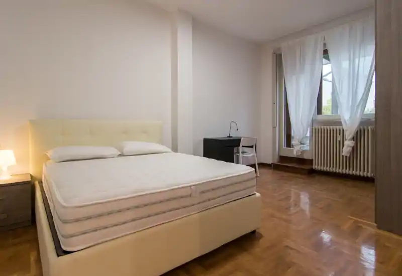 Bright, modern bedroom with a large upholstered bed, bedside table and lamp, desk by the window, and polished parquet flooring — ideal for rental photos.
