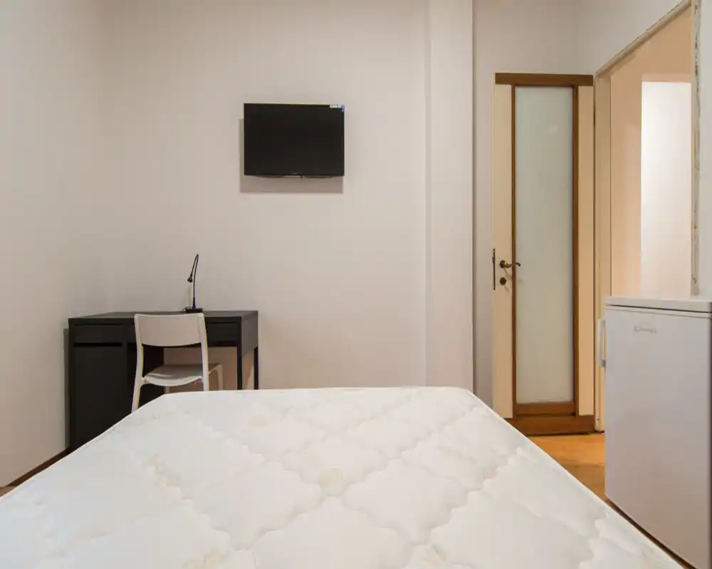 Camera da letto semplice e ben illuminata con un grande materasso in primo piano, TV a parete e una scrivania con sedia — ideale per un viaggiatore singolo o soggiorni brevi.