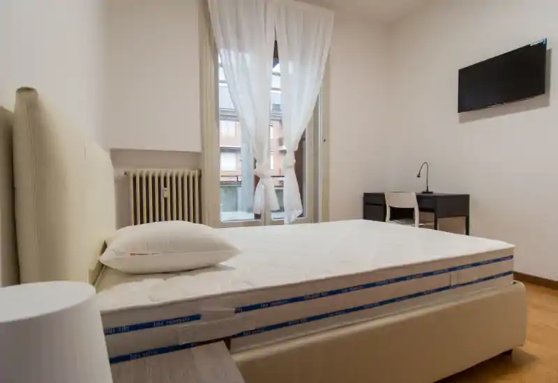 Camera da letto luminosa e moderna con un grande letto imbottito, cuscino e luce naturale da una finestra con tende leggere; scrivania semplice e TV a parete sullo sfondo.