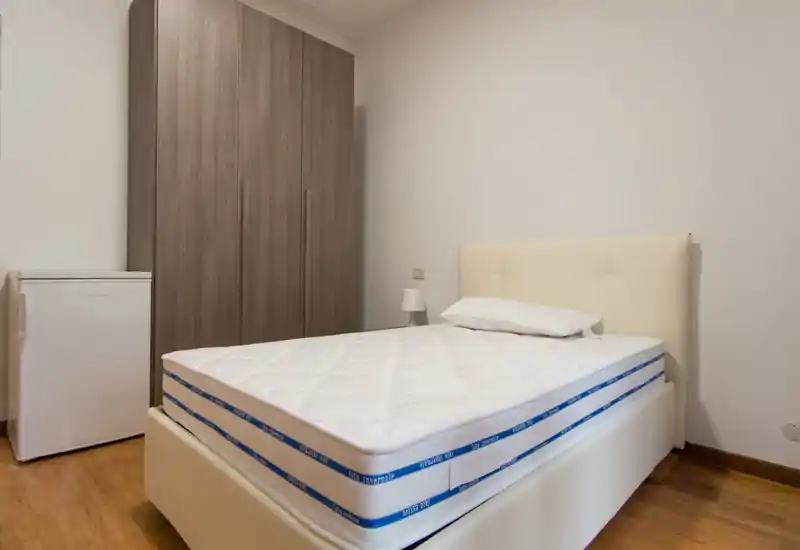 Camera da letto ordinata con letto imbottito, materasso, lampada da comodino e grande armadio — arredi neutri e luminosi ideali per annunci.