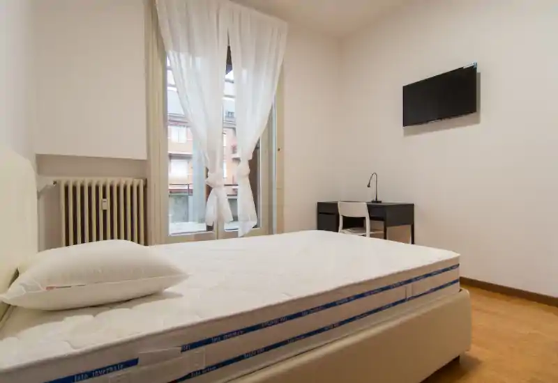 Camera da letto luminosa e minimal con materasso ben visibile in primo piano, biancheria bianca, scrivania e TV a parete — ideale per soggiorni singoli.