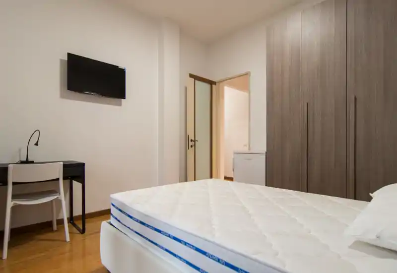 Camera da letto luminosa e ordinata con un grande materasso in primo piano, un armadio a parete e una piccola scrivania con TV a muro — allestimento minimal e moderno ideale per affitto.