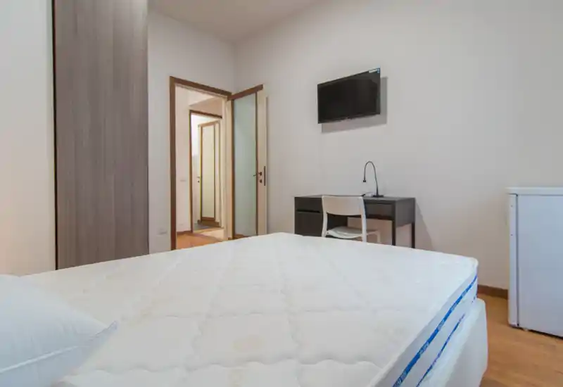 Dormitorio moderno y luminoso con un gran colchón en primer plano, TV de pared, escritorio y armario; estilo limpio y minimalista ideal para anuncios.