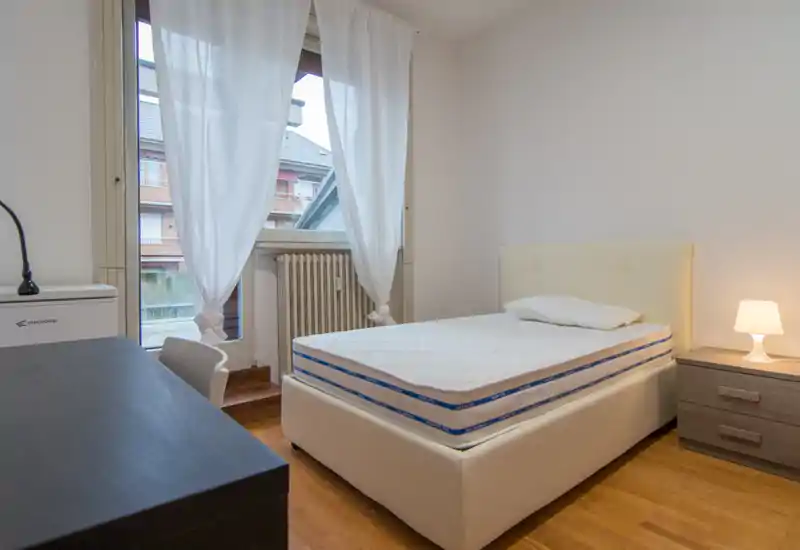 Dormitorio luminoso y de mobiliario sencillo con cama individual, mesita y escritorio junto a una gran ventana con cortinas translúcidas, ideal para alquileres compactos.