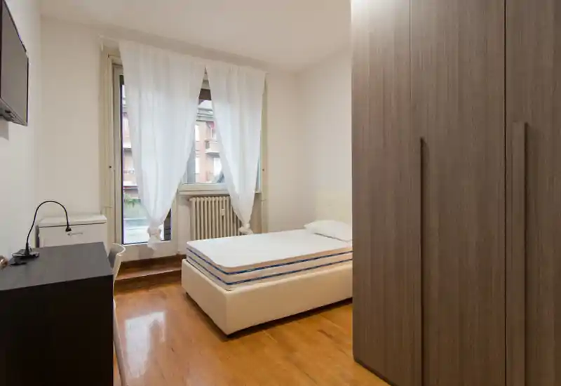 Dormitorio luminoso y de mobiliario sencillo con cama individual, armario y escritorio. La luz natural de la ventana genera un ambiente limpio y acogedor.