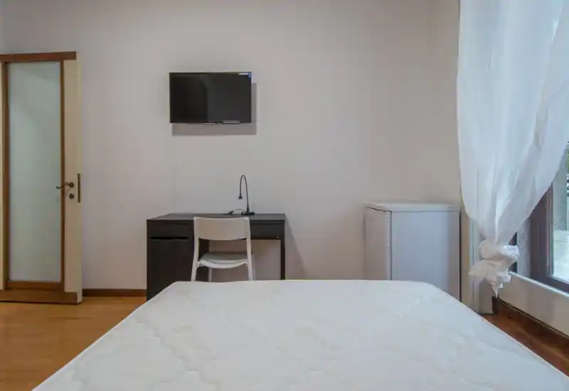 Camera da letto semplice e luminosa con materasso in primo piano, piccolo scrittoio e sedia contro la parete, TV a muro e finestra con tende leggere che lasciano entrare luce naturale.