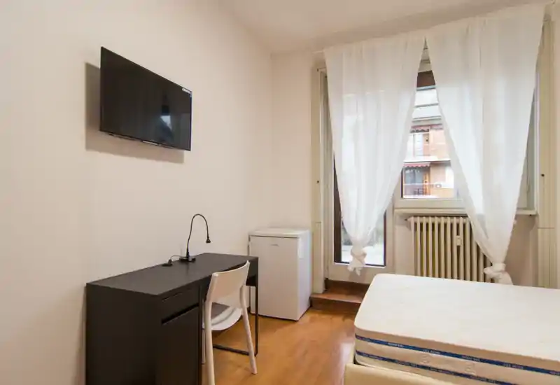Camera da letto luminosa e arredata in modo semplice con materasso visibile, scrivania, TV a parete e luce naturale da una grande finestra con tende leggere.