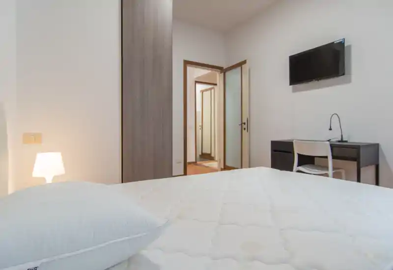 Dormitorio luminoso y minimalista con un gran colchón visible, lámpara de mesilla y un pequeño escritorio con TV en la pared — amplio y de mobiliario sencillo.