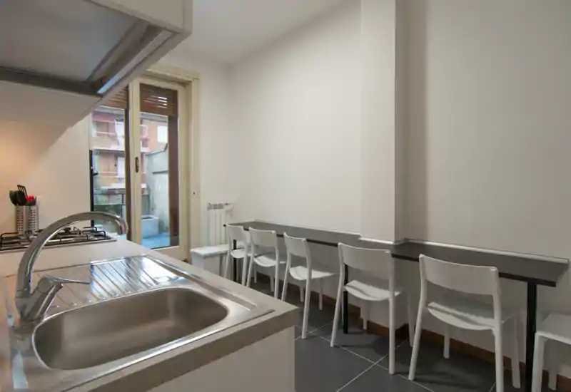 Bright, compact kitchen with stainless sink, gas hob and a long counter with chairs facing the wall; natural light comes from a balcony door, giving a practical layout ideal for a small apartment.