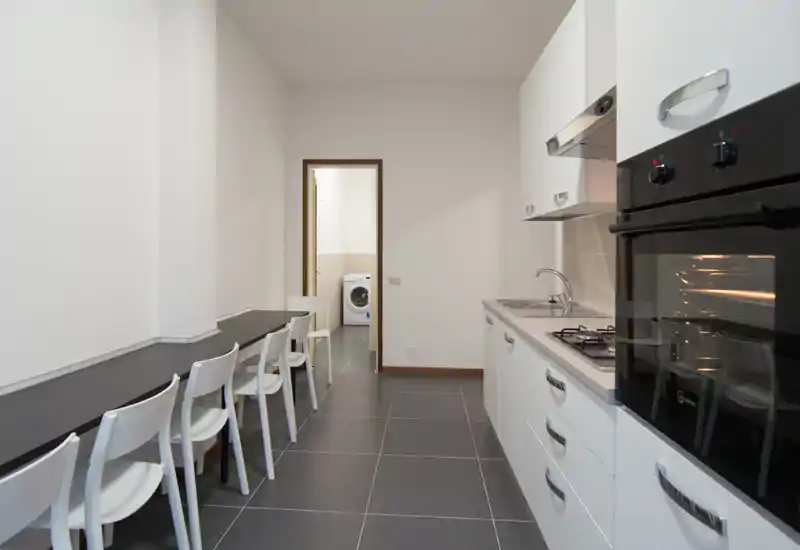Cocina moderna en línea con muebles blancos, horno empotrado, placa de gas y una larga barra con sillas — distribución limpia y funcional, ideal para apartamentos compactos.