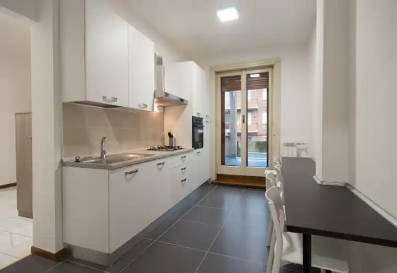 Modern galley kitchen with white cabinets, integrated oven and gas hob, a stainless sink and a narrow breakfast counter with stools. Bright and neat space with a door opening to a small balcony.
