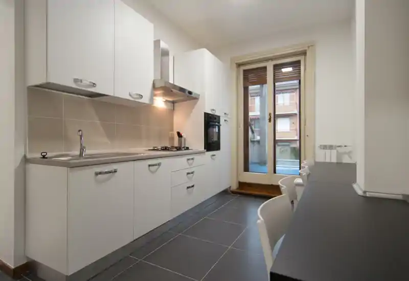 Bright modern galley kitchen with white cabinets, integrated oven and gas hob, a sink on the left and a breakfast counter with stools on the right. Large glass door provides natural light and access to a small balcony.