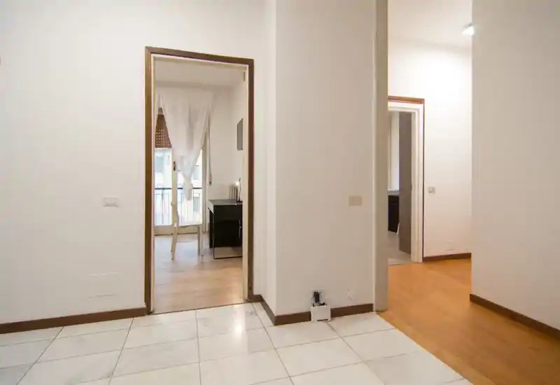 Interno luminoso e minimale che mostra un ingresso/corridoio con aperture verso stanze adiacenti; pavimento in piastrelle in primo piano e legno oltre, con luce naturale proveniente da una stanza con balcone.