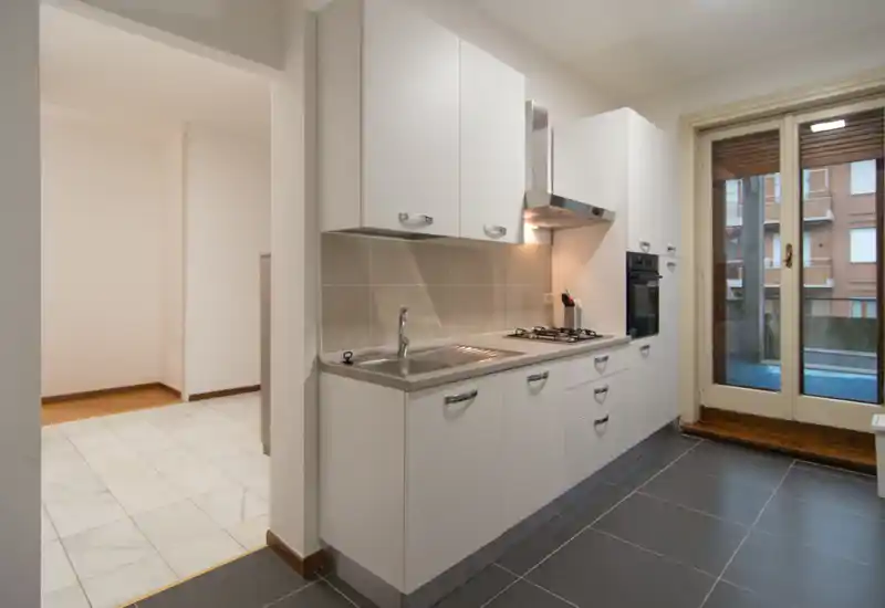 Bright, modern fitted kitchen with white cabinets, gas hob, built-in oven and sink; large window/door to a small balcony provides natural light.