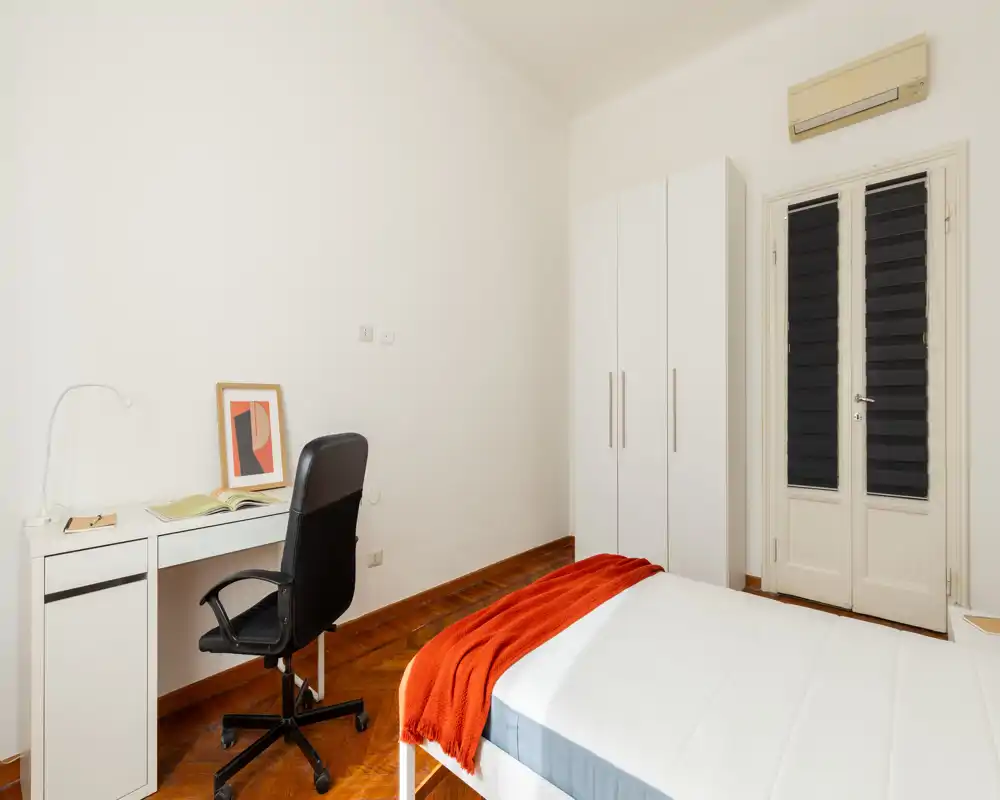 Camera da letto luminosa e minimal con letto rifatto, armadio bianco e una scrivania compatta con sedia da ufficio — stile pulito e plaid arancione che aggiunge calore.