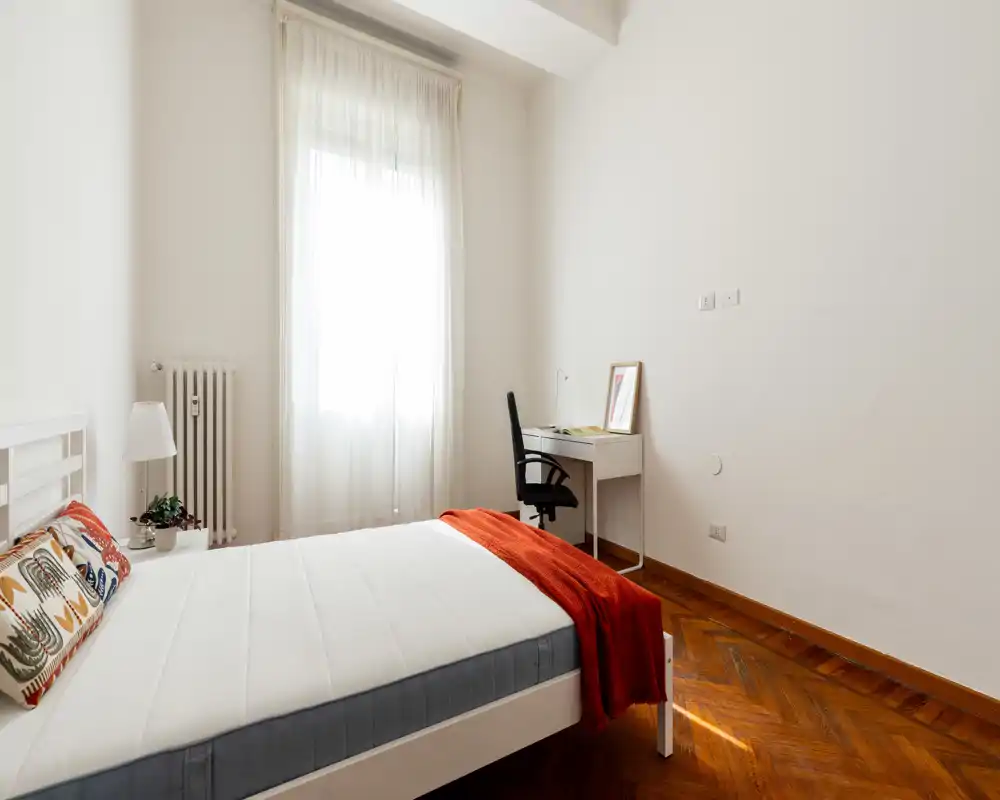 Camera da letto luminosa e minimal con letto bianco, cuscini decorativi e un plaid rosso; una scrivania con sedia si trova vicino a una finestra alta che diffonde luce naturale.