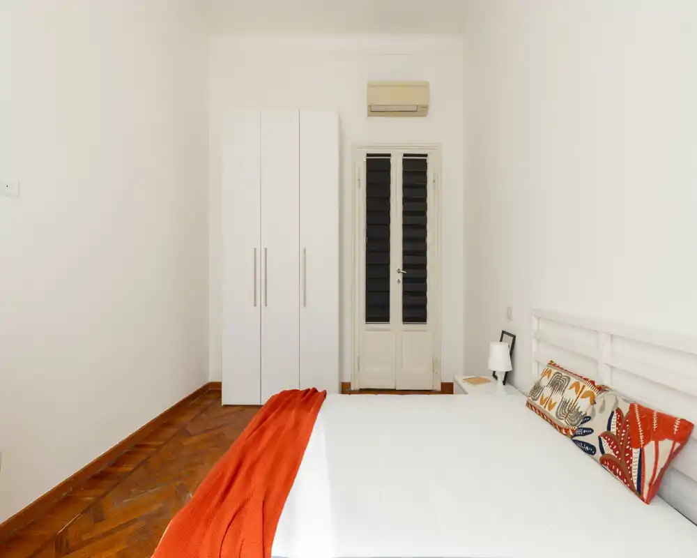 Camera da letto luminosa e minimal con un grande letto bianco, cuscini colorati e un plaid arancione. L'ambiente include un armadio bianco, pavimento in parquet e un comodino con lampada — perfetto per un annuncio di affitto moderno.