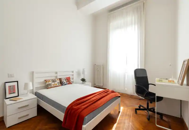 Camera da letto luminosa e minimal con letto in legno bianco, cuscini decorativi e una coperta arancione; ampia finestra con molta luce naturale e una postazione con sedia da ufficio.