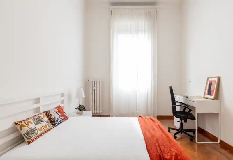Camera da letto luminosa e minimal con ampio letto bianco, cuscini decorativi e un plaid arancione; una scrivania con sedia vicino alla finestra offre un comodo spazio di lavoro.