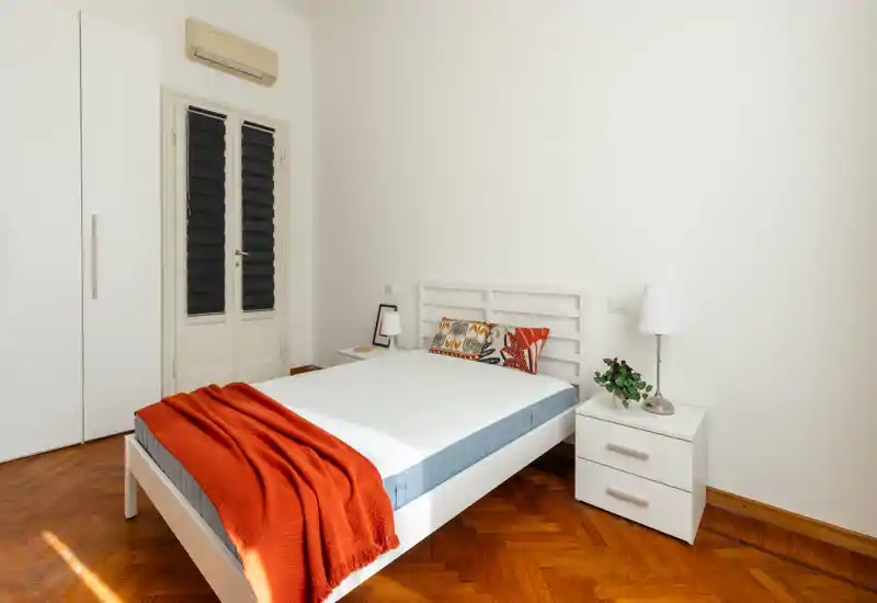 Camera da letto luminosa e minimal con struttura del letto bianca, cuscini colorati e un plaid arancione su pavimento in parquet. Comodino con lampada e armadio completano l’ambiente, perfetto per annunci di affitto.