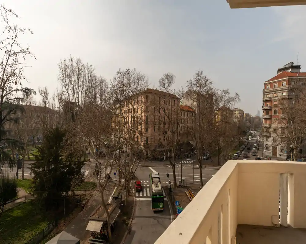 Balcón soleado con vistas a una calle urbana arbolada, edificios de altura media y una parada de tranvía/autobús abajo.