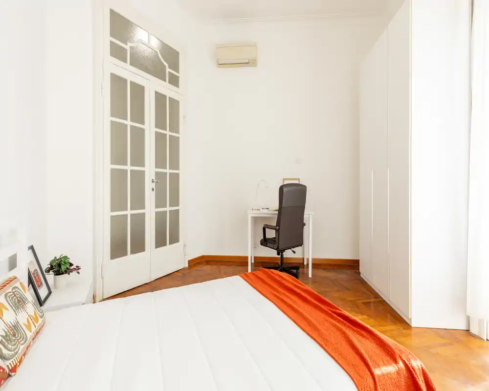 Dormitorio luminoso y minimalista con una cama grande en primer plano, suelo de parquet, un armario blanco y un pequeño espacio de trabajo junto a la pared. La habitación es luminosa y ventilada, con un acento cálido con la manta naranja.