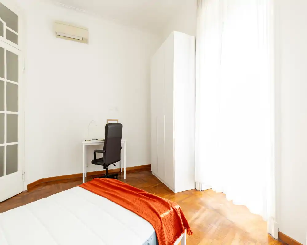 Dormitorio luminoso y minimalista con ventana grande, paredes blancas, armario y cama bien hecha con manta naranja; un pequeño escritorio y silla ofrecen un rincón de trabajo.
