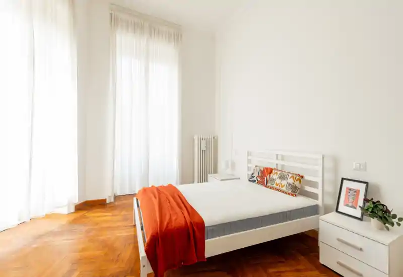 Dormitorio luminoso y minimalista con cama blanca, cojines coloridos y una manta naranja sobre parquet de madera cálida. Grandes ventanas con cortinas ligeras proporcionan abundante luz natural y un ambiente tranquilo y acogedor.